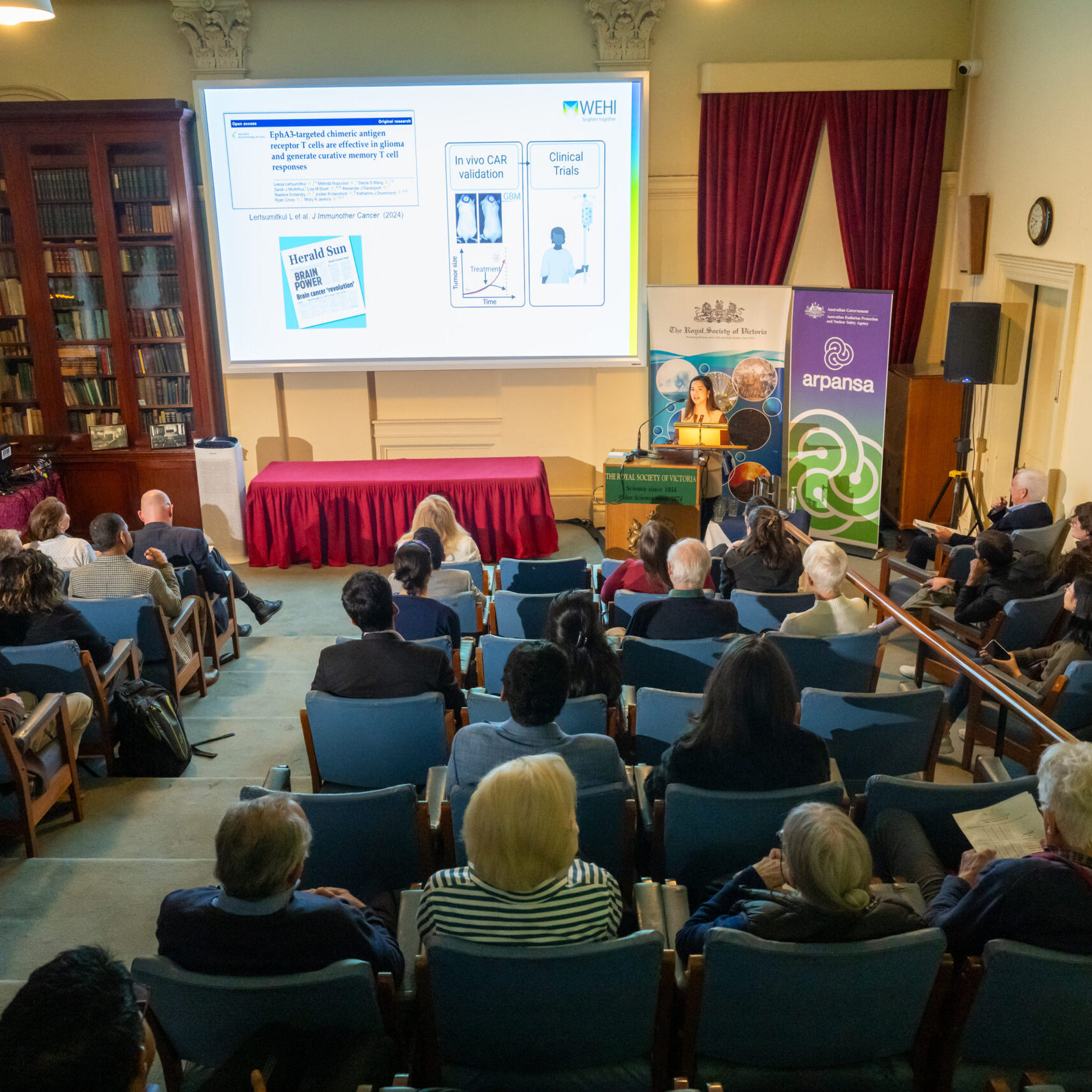 An audience in a lecture theatre listening to a presentation from a person at the lectern with images projected on a screen