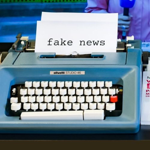 Typewriter with a page reading "fake news" in the carriage. A television displaying a news report is in the background.